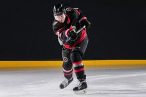 Hockey player in a black and red uniform skating forward on an indoor ice rink, gripping a stick as snow-like ice chips fly up.