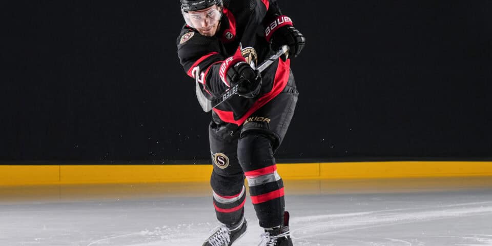 Hockey player in a black and red uniform skating forward on an indoor ice rink, gripping a stick as snow-like ice chips fly up.