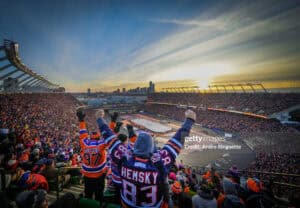 Fans in orange and blue jerseys cheer from the packed hockey arena as the sunset lights the city skyline in the background.