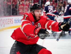 Canadian hockey player in a red jersey and helmet, skating during a game with spectators in the stands behind him.