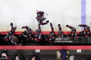 Ice hockey players in black and red uniforms celebrate along the boards, one player leaping mid-air while teammates raise their fists in celebration.