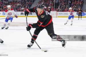 Hockey player in a black and red jersey controls the puck during a fast-paced game on the ice online with a crowd in the background.