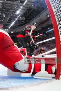 Hockey goalie in red-and-black jersey (#19) stands by the crease as the arena fills with spectators.