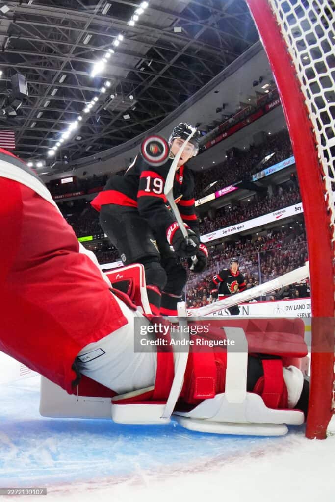 1 2026 459 | Photography and video news, reviews and tips | Henry's Camera Hockey goalie in red-and-black jersey (#19) stands by the crease as the arena fills with spectators.