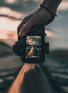 21camera | Photography and video news, reviews and tips | Henry's Camera Hand holding a DSLR aimed along a railroad track toward a sunset horizon, the camera screen showing the scene