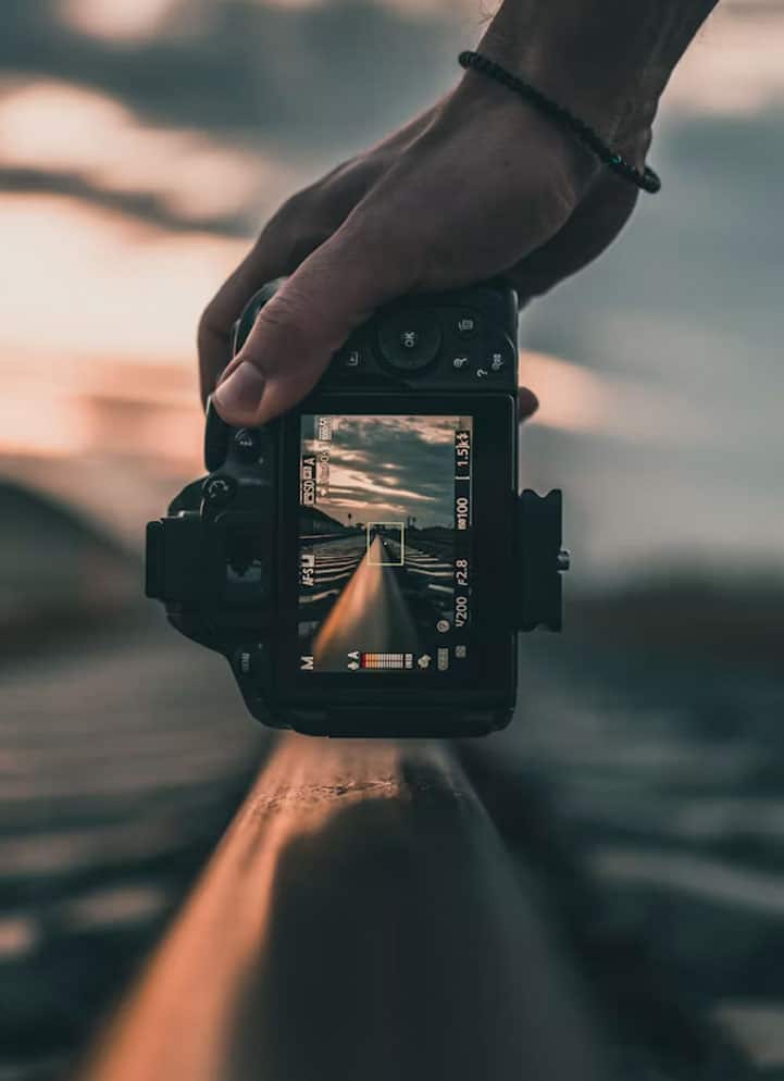 Hand holding a DSLR aimed along a railroad track toward a sunset horizon, the camera screen showing the scene