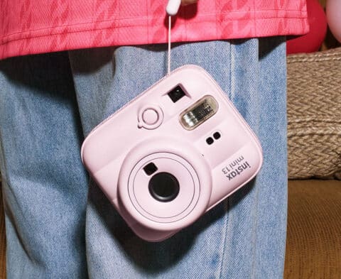 Pink Instax Mini 13 instant camera hanging from a strap against a person in a pink jacket and blue jeans.