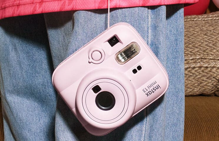 Pink Instax Mini 13 instant camera hanging from a strap against a person in a pink jacket and blue jeans.
