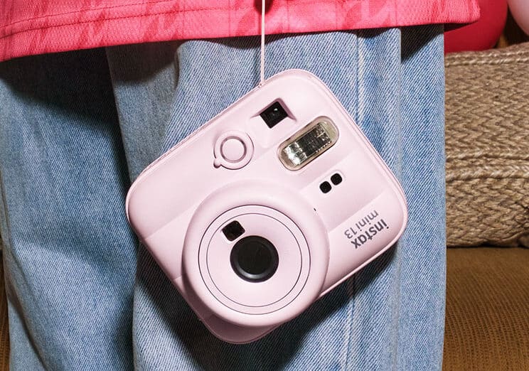 3Instax | Photography and video news, reviews and tips | Henry's Camera Pink Instax Mini 13 instant camera hanging from a strap against a person in a pink jacket and blue jeans.