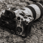 Canon EOS R5 DSLR with a cracked LCD screen resting on a rocky surface, camo-patterned body and attached telephoto lens present.