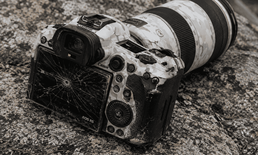Canon EOS R5 DSLR with a cracked LCD screen resting on a rocky surface, camo-patterned body and attached telephoto lens present.