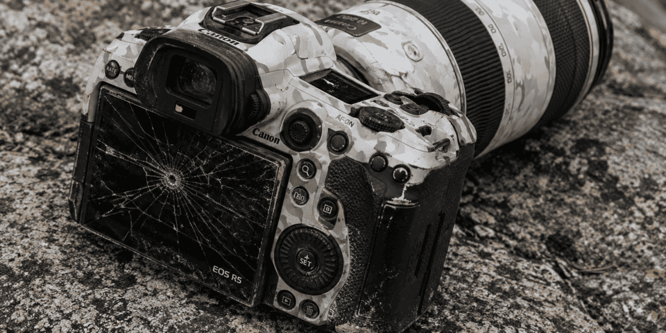 Canon EOS R5 DSLR with a cracked LCD screen resting on a rocky surface, camo-patterned body and attached telephoto lens present.