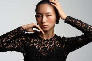 Portrait of a woman in a black lace top, hands in her hair, neutral expression against a gray backdrop.