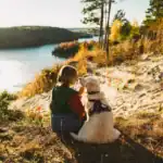 Woman in a red jacket and green vest sits on a sandy hill with a golden retriever, facing a lake surrounded by autumn trees.