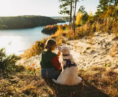 Woman in a red jacket and green vest sits on a sandy hill with a golden retriever, facing a lake surrounded by autumn trees.