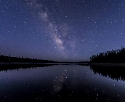 Star-filled night sky with the Milky Way over a calm lake, reflected in the water, with a dark forest along the shore.