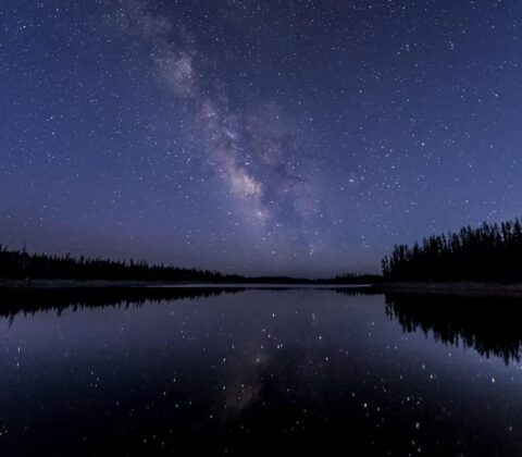 Star-filled night sky with the Milky Way over a calm lake, reflected in the water, with a dark forest along the shore.