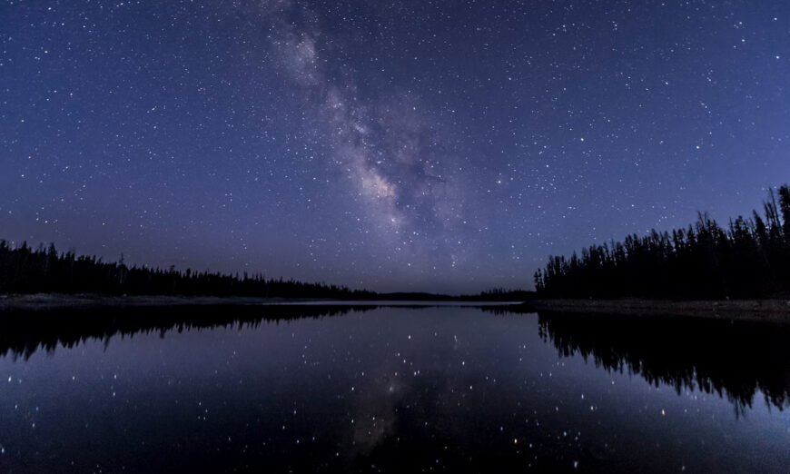 Astro-1 | Photography and video news, reviews and tips | Henry's Camera Star-filled night sky with the Milky Way over a calm lake, reflected in the water, with a dark forest along the shore.