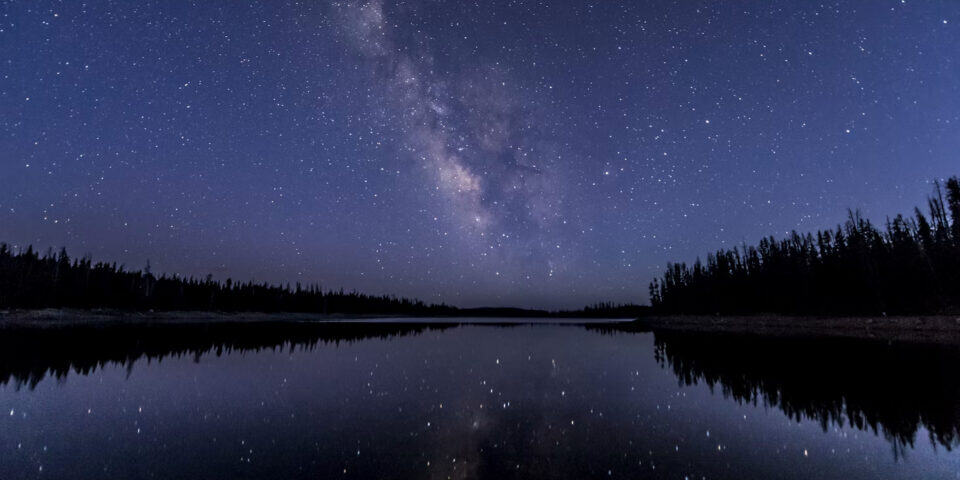 Star-filled night sky with the Milky Way over a calm lake, reflected in the water, with a dark forest along the shore.