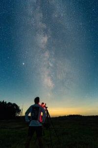 Person setting up a telescope under a star-filled night sky with the Milky Way visible above a horizon at dusk.