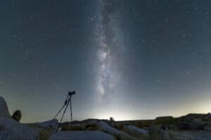 Milky Way stretches across a starry night sky above a rocky desert, with a camera on a tripod pointed upward in the foreground.