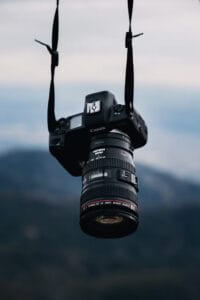 Canon EOS DSLR camera with lens hanging from straps against a blurred mountain landscape in the background