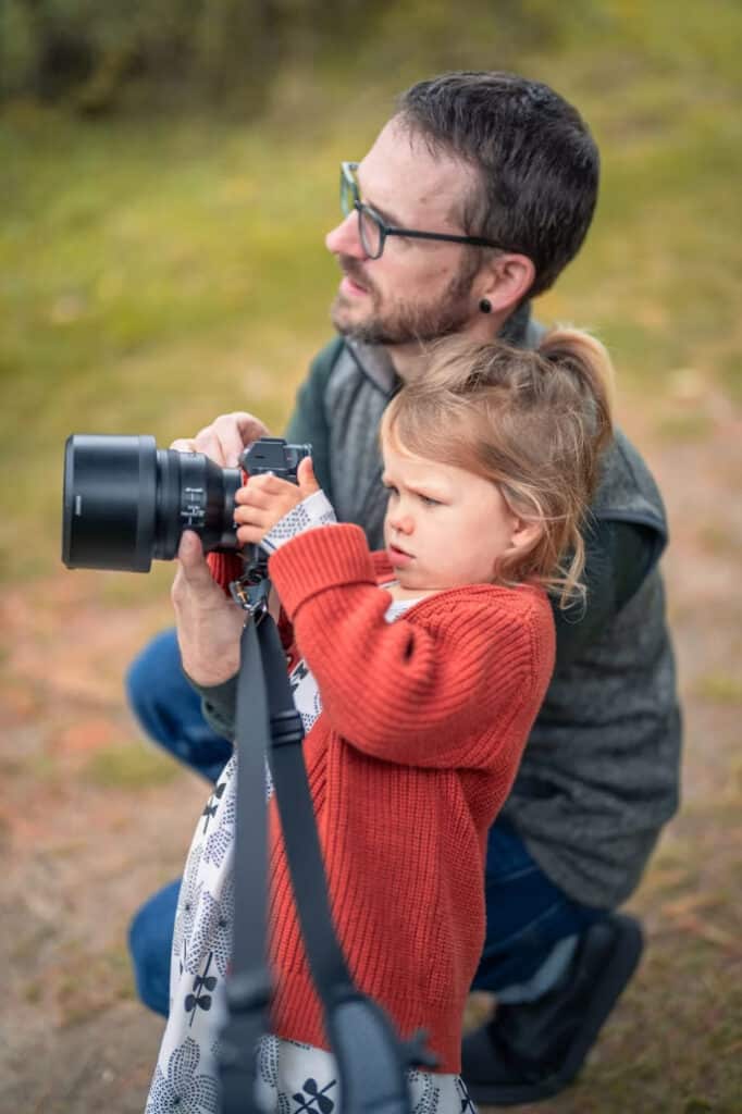 Dad | Photography and video news, reviews and tips | Henry's Camera Father kneels beside daughter as she holds a DSLR with a large lens outdoors, learning photography.