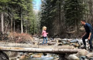 Father and young daughter balance across a fallen log over a rocky stream in a forest.