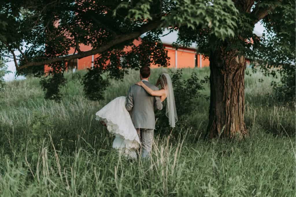 Bride in a white gown and veil is carried by the groom as they walk away through tall grass under a large tree, with a red barn in the background.
