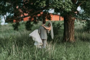 Bride in a white gown and veil is carried by the groom as they walk away through tall grass under a large tree, with a red barn in the background.