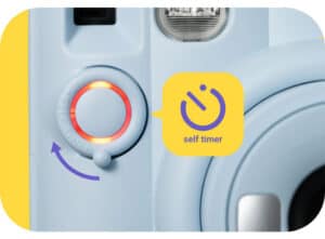 Close-up of a light-blue camera with a red-ringed shutter button, purple arrow, and a yellow speech bubble that says 'self timer'.