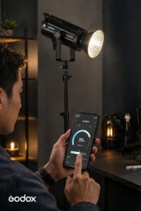 Person adjusts a Godox SL200II studio light using a smartphone app at 70% brightness on a dimly lit set.