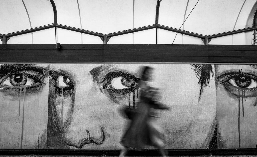 Large mural of a crying pair of eyes with dripping tears, a blurred passerby walking past.