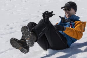 Guy-sliding | Photography and video news, reviews and tips | Henry's Camera Smiling man in a blue and orange jacket lying in the snow, holding a handheld camera.