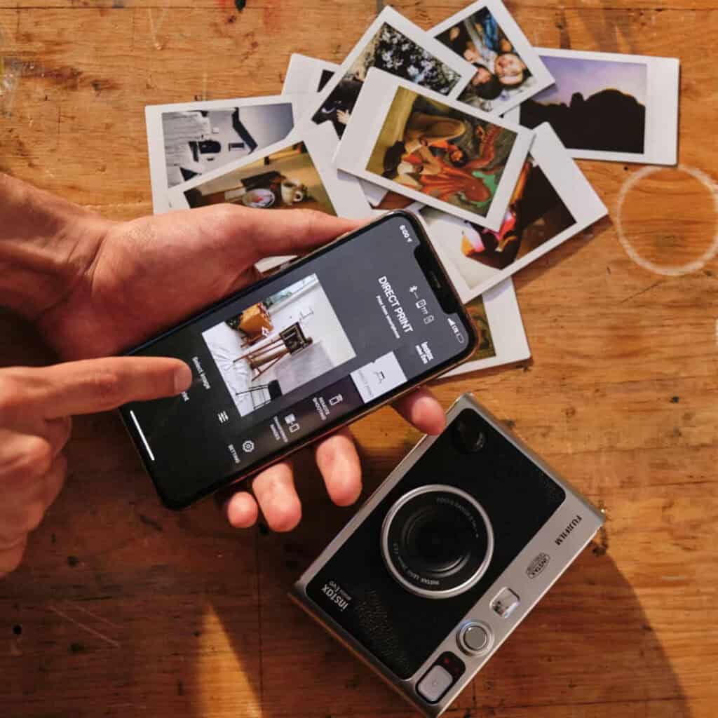 InstaxEvo2 | Photography and video news, reviews and tips | Henry's Camera Hands hold a smartphone showing a photo-printing app while scattered instant photos lie on a wooden table beside a compact camera.
