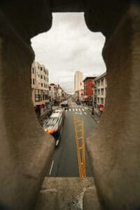 View of a busy city street framed by stone balusters, showing buildings, crosswalks, and moving traffic from a high vantage point in a narrow opening.