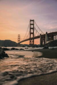 KN407801 | Photography and video news, reviews and tips | Henry's Camera Golden Gate Bridge at sunset viewed from a rocky beach with calm water in the foreground.