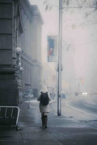 Person walking away on a snow-dusted city sidewalk, wearing a beige coat and backpack, with blurred buildings and streetlamp in the background