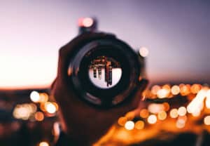 Close-up of a camera lens held up to the scene, with warm city lights blurred in the background and the skyline reflected upside down in the lens.