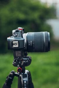 Canon DSLR with a large zoom lens mounted on a tripod outdoors, blurred green background behind.