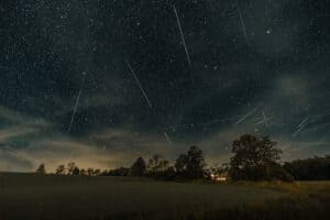 Night sky full of stars and several meteor streaks over a field with trees and a lit house in the distance at night.