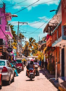 Colorful tropical street scene with pedestrians, motorbikes, and cars on a cobblestone road; bright pink and orange buildings with palm trees under a blue sky.