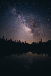 Night sky filled with stars and the Milky Way above a dark forest reflected in a calm lake in the foreground.