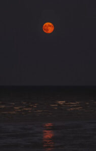 Bright orange full moon over dark sea with its red reflection shimmering on gentle waves at night.