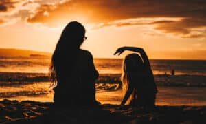 Two people sit on a sandy beach at sunset, silhouettes against the orange sky, one raising an arm gracefully.