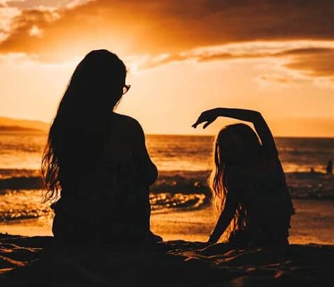 Two people sit on a sandy beach at sunset, silhouettes against the orange sky, one raising an arm gracefully.