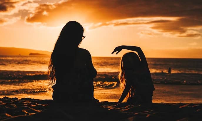MothersDay | Photography and video news, reviews and tips | Henry's Camera Two people sit on a sandy beach at sunset, silhouettes against the orange sky, one raising an arm gracefully.