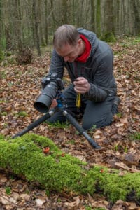 Photographer kneeling in a leafy forest, adjusting a DSLR on a small tripod over mossy ground.