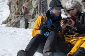 Two photographers sit on snow beside a rocky cliff, comparing gear and cameras in cold weather.