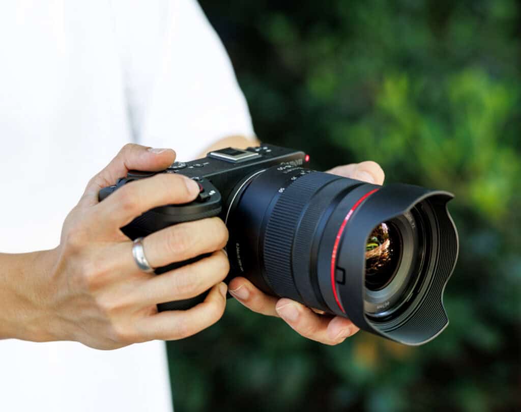 Person holding a black camera with a large lens outdoors, as if ready to shoot a photo.